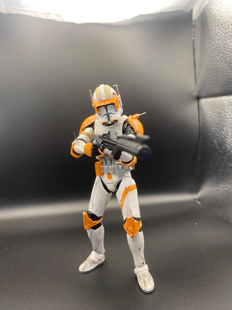 Pu&ograve; includere: Una action figure bianca e arancione di un clone trooper, che tiene in mano un fucile blaster nero. La figura ha un elmetto con segni arancioni e dettagli dell'armatura. La figura &egrave; in posizione di combattimento.