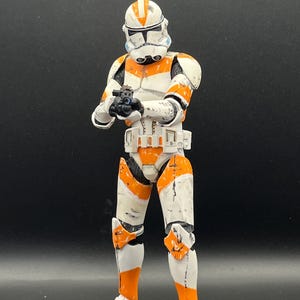 Pu&ograve; includere: Action figure dettagliata di un clone trooper in armatura bianca e arancione, con un blaster. La figura ha un elmetto con segni arancioni ed &egrave; posizionata su uno sfondo scuro. La figura &egrave; alta circa 15 cm.
