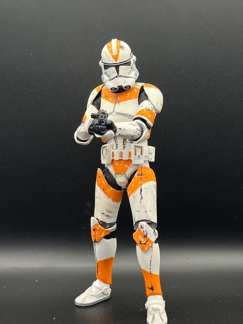 Pu&ograve; includere: Action figure dettagliata di un clone trooper in armatura bianca e arancione, con un blaster. La figura ha un elmetto con segni arancioni ed &egrave; posizionata su uno sfondo scuro. La figura &egrave; alta circa 15 cm.