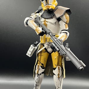 Pu&ograve; includere: Action figure dettagliata di un clone trooper in armatura bianca e gialla, con un fucile blaster. La figura ha un elmetto dal design distintivo ed &egrave; posata su uno sfondo scuro. L'armatura mostra segni di usura.