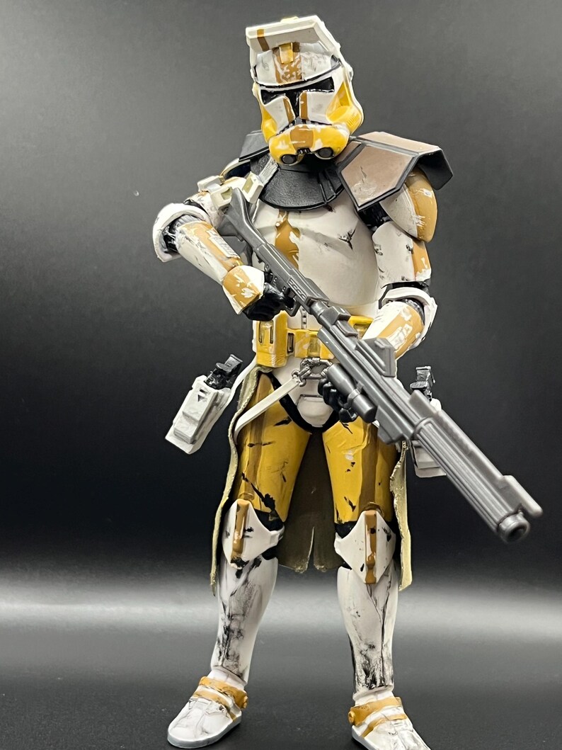 Pu&ograve; includere: Action figure dettagliata di un clone trooper in armatura bianca e gialla, con un fucile blaster. La figura ha un elmetto dal design distintivo ed &egrave; posata su uno sfondo scuro. L'armatura mostra segni di usura.