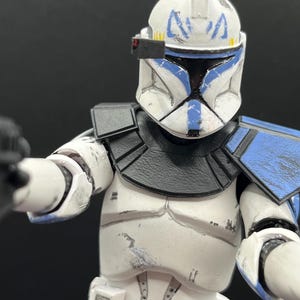 Pu&ograve; includere: Una action figure bianca e blu di un clone trooper di un franchise di fantascienza. La figura ha un elmetto con segni blu, una visiera nera e un corpo bianco con dettagli di armatura blu e neri. La figura tiene in mano un'arma nera.