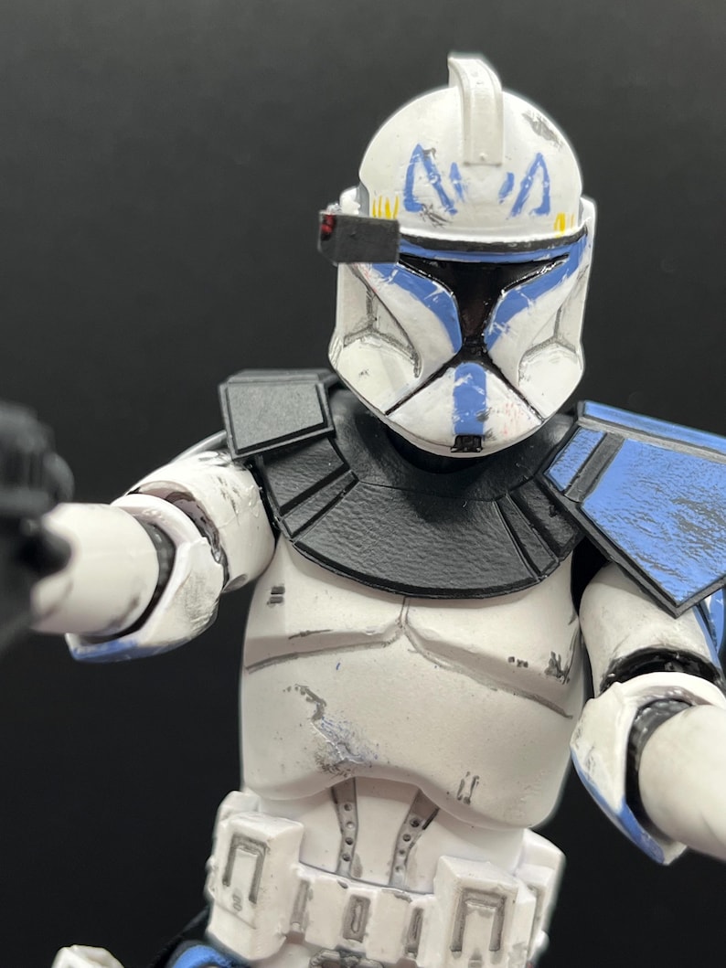 Pu&ograve; includere: Una action figure bianca e blu di un clone trooper di un franchise di fantascienza. La figura ha un elmetto con segni blu, una visiera nera e un corpo bianco con dettagli di armatura blu e neri. La figura tiene in mano un'arma nera.