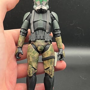 Pu&ograve; includere: Action figure dettagliata di un soldato in uniforme verde e mimetica. La figura ha un elmetto con dettagli verdi, un pettorale nero e pantaloni mimetici. La figura &egrave; tenuta in una mano.