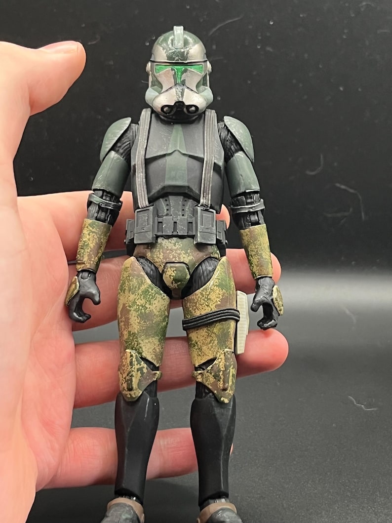 Pu&ograve; includere: Action figure dettagliata di un soldato in uniforme verde e mimetica. La figura ha un elmetto con dettagli verdi, un pettorale nero e pantaloni mimetici. La figura &egrave; tenuta in una mano.