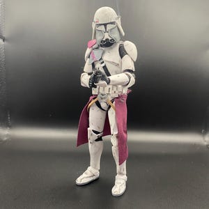 Pu&ograve; includere: Action figure bianca e grigia di un soldato in armatura, con un'arma nera. La figura ha un elmetto, spalline e una sciarpa bordeaux. La figura &egrave; su una superficie scura.