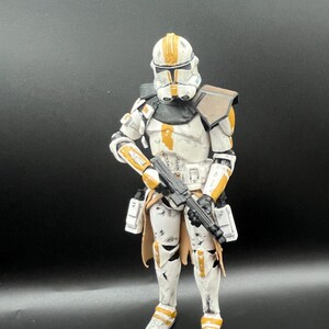 Pu&ograve; includere: Una dettagliata action figure di un clone trooper di Star Wars. La figura &egrave; prevalentemente bianca con dettagli arancioni e neri. Tiene in mano un fucile blaster nero ed &egrave; posizionata su uno sfondo scuro.