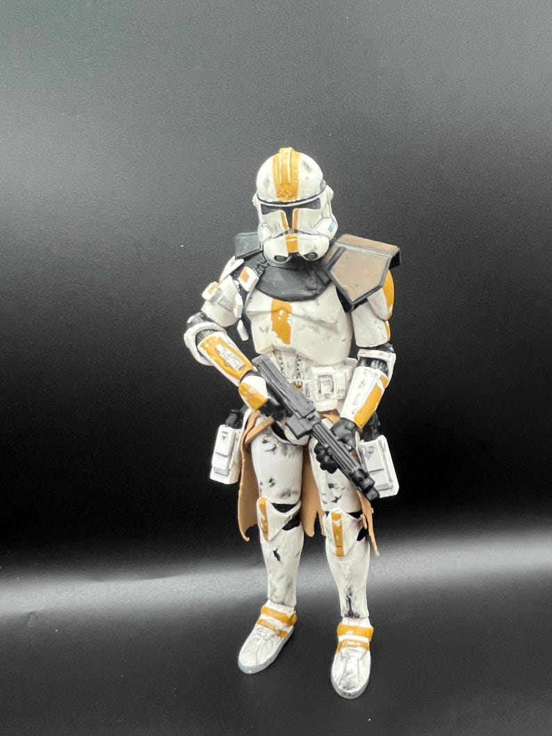 Pu&ograve; includere: Una dettagliata action figure di un clone trooper di Star Wars. La figura &egrave; prevalentemente bianca con dettagli arancioni e neri. Tiene in mano un fucile blaster nero ed &egrave; posizionata su uno sfondo scuro.