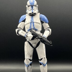 Pu&ograve; includere: Action figure bianca e blu di un clone trooper di un franchise di fantascienza. La figura &egrave; in posizione eretta, con un fucile blaster nero. L'armatura ha accenti blu sul casco, sulle spalle e sulle ginocchia. Su sfondo nero.