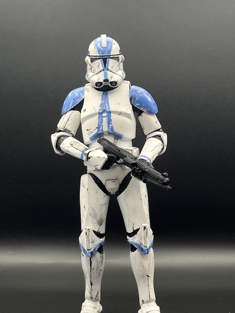 Pu&ograve; includere: Action figure bianca e blu di un clone trooper di un franchise di fantascienza. La figura &egrave; in posizione eretta, con un fucile blaster nero. L'armatura ha accenti blu sul casco, sulle spalle e sulle ginocchia. Su sfondo nero.