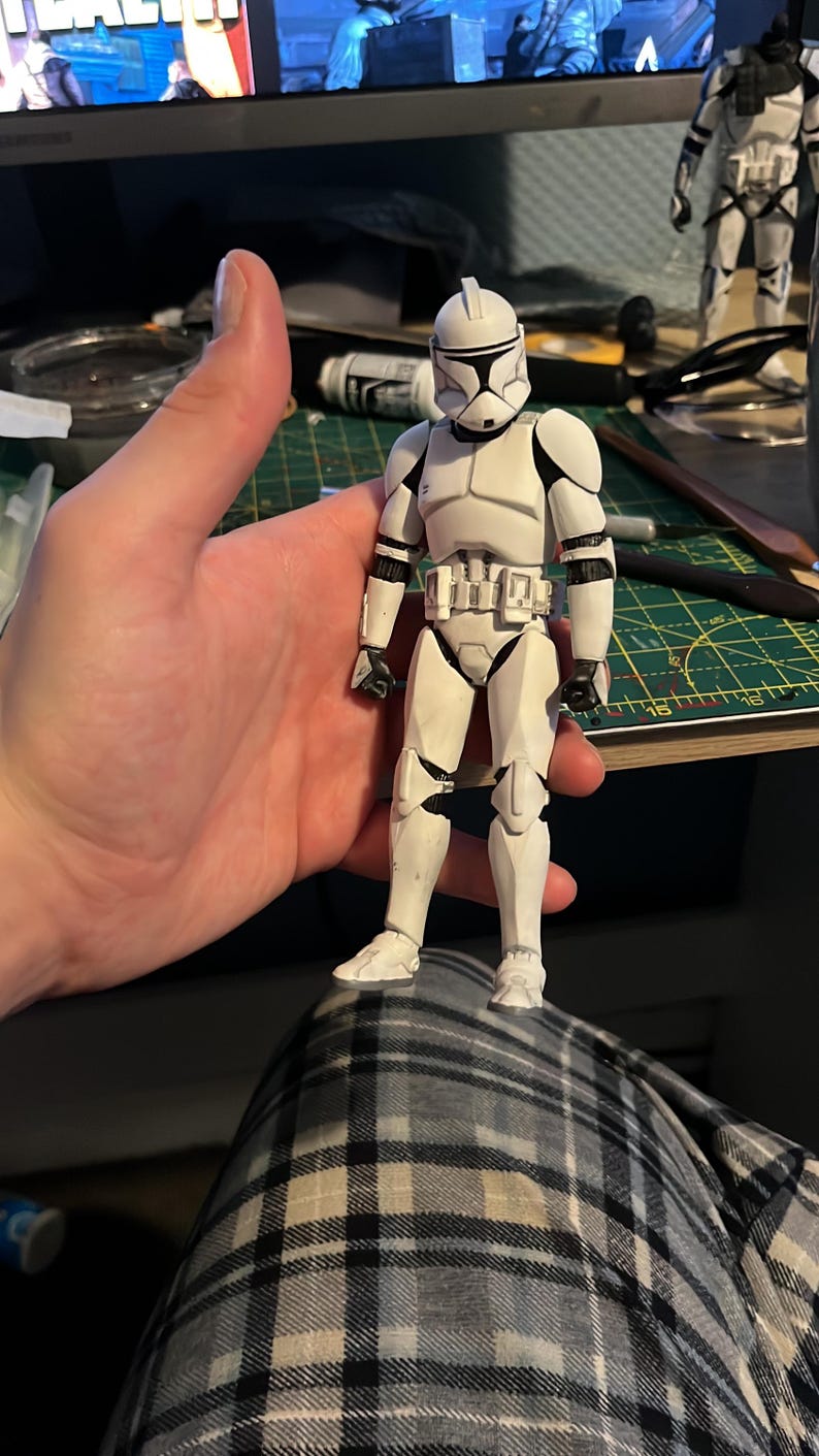 Pu&ograve; includere: Action figure bianca e nera di un clone trooper di Star Wars. La figura &egrave; tenuta in mano da una persona e indossa un elmetto e un'armatura bianchi. La figura &egrave; alta circa 15 cm.