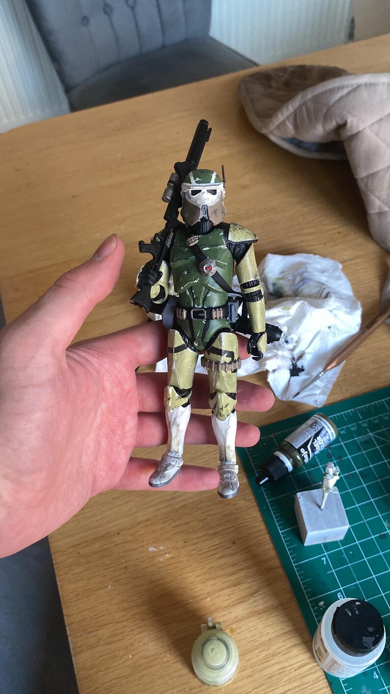Pu&ograve; includere: Action figure dettagliata di un soldato con armatura verde oliva e beige, che tiene un fucile nero. La figura ha un elmetto bianco ed &egrave; posta su uno sfondo neutro. La figura &egrave; alta circa 15 cm.