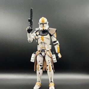 Pu&ograve; includere: Una action figure bianca di un clone trooper con dettagli arancioni e marroni, che tiene in mano un fucile blaster nero. La figura ha un'armatura dettagliata e si trova su uno sfondo scuro. Il casco ha un design distintivo.