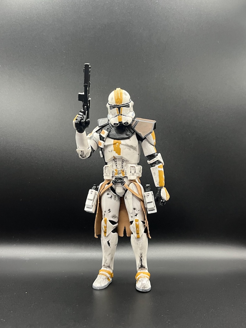 Pu&ograve; includere: Una action figure bianca di un clone trooper con dettagli arancioni e marroni, che tiene in mano un fucile blaster nero. La figura ha un'armatura dettagliata e si trova su uno sfondo scuro. Il casco ha un design distintivo.