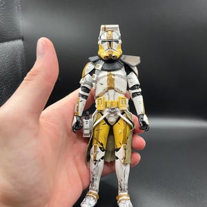 Pu&ograve; includere: Action figure dettagliata di un clone trooper di Star Wars. La figura &egrave; principalmente bianca con accenti gialli e marroni, tra cui il casco, il petto e le gambe. Ha dettagli neri e tiene un piccolo accessorio rettangolare.