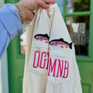 May include: A cream-colored long-sleeve shirt featuring two chest pockets. Each pocket is embroidered with a colorful trout fish above large, bright pink block-letter monograms reading OGM and MNB in a vertical, stacked font style.