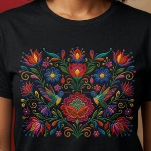 May include: A black t-shirt featuring a vibrant, multi-colored embroidered-style graphic on the chest. The design displays symmetrical floral patterns, swirling vines, and two hummingbirds in shades of red, blue, green, yellow, and pink.
