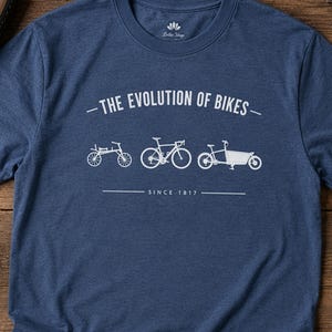 Può includere: Una t-shirt a maniche corte blu navy mélange con una stampa grafica bianca. Il design mostra tre sagome di biciclette con il testo "THE EVOLUTION OF BIKES" sopra e "SINCE 1817" sotto. La maglietta ha un girocollo e un logo del marchio.