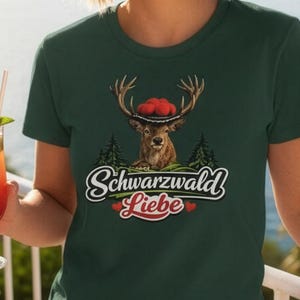 May include: A dark green t-shirt featuring a graphic of a deer wearing a traditional hat with red pom-poms, surrounded by pine trees. Below the deer, the text reads "Schwarzwald Liebe" in a white script font, accented by two small red hearts.
