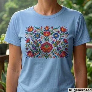 May include: A light blue short-sleeved t-shirt featuring a central rectangular graphic print. The design displays a symmetrical arrangement of colorful flowers, swirling green vines, and two hummingbirds facing each other in vibrant hues.