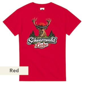 May include: A bright red short-sleeved t-shirt featuring a graphic of a deer wearing a traditional red pom-pom hat. Below the deer are green pine trees and the text Schwarzwald Liebe in a white script font with a black outline and red hearts.