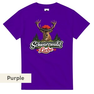 May include: A purple short-sleeved t-shirt featuring a graphic of a deer wearing a red pom-pom hat. Below the deer are green pine trees and the text Schwarzwald Liebe in white script with two small red hearts on either side of the word Liebe.