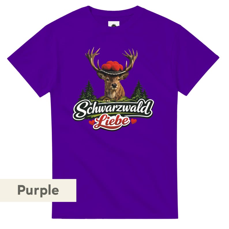May include: A purple short-sleeved t-shirt featuring a graphic of a deer wearing a red pom-pom hat. Below the deer are green pine trees and the text Schwarzwald Liebe in white script with two small red hearts on either side of the word Liebe.