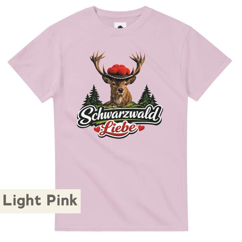 May include: A light pink short-sleeve t-shirt featuring a graphic of a deer wearing a traditional hat with red pom-poms, set against a backdrop of pine trees and a small house, with the text Schwarzwald Liebe and two red hearts below.
