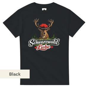 May include: A black short-sleeved t-shirt featuring a graphic of a deer wearing a traditional hat with red pom-poms. Below the deer are green pine trees and the white text Schwarzwald Liebe with two small red hearts on either side of the word Liebe.