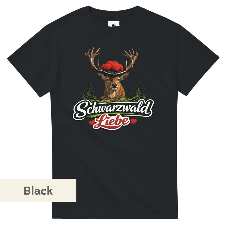 May include: A black short-sleeved t-shirt featuring a graphic of a deer wearing a traditional hat with red pom-poms. Below the deer are green pine trees and the white text Schwarzwald Liebe with two small red hearts on either side of the word Liebe.
