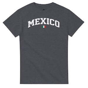 Puede incluir: Camiseta gris oscuro con la palabra "MEXICO" en blanco, con letras arqueadas. Una peque&ntilde;a bandera mexicana est&aacute; centrada debajo del texto. La camiseta es de cuello redondo.