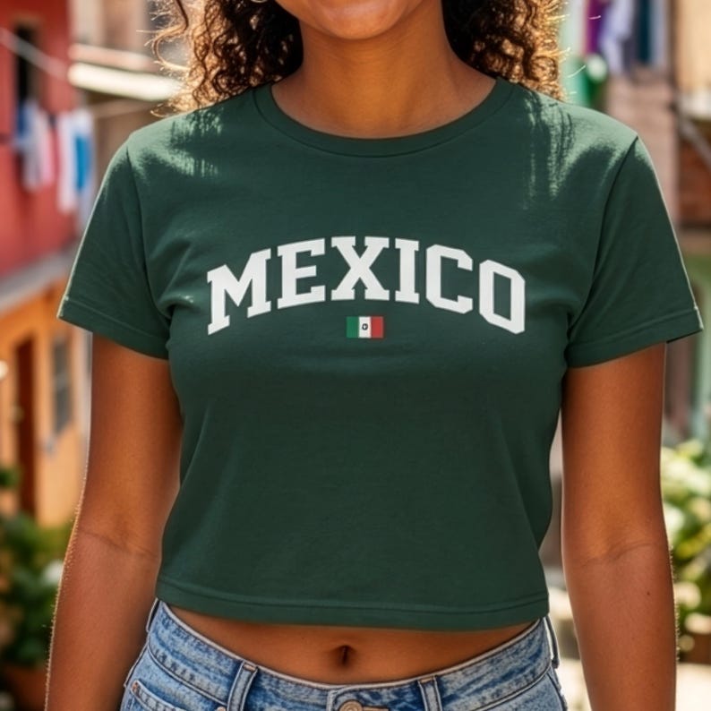 Puede incluir: Una camiseta corta verde oscuro con la palabra "MEXICO" en letras blancas. Debajo del texto hay una peque&ntilde;a bandera mexicana. La camiseta est&aacute; hecha de un material suave y se combina con vaqueros azules.