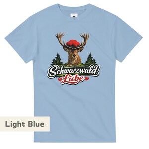 May include: A light blue short-sleeve t-shirt featuring a graphic of a deer wearing a traditional hat with red pom-poms, set against a forest background. Below the deer, the text reads Schwarzwald Liebe with two small red hearts.