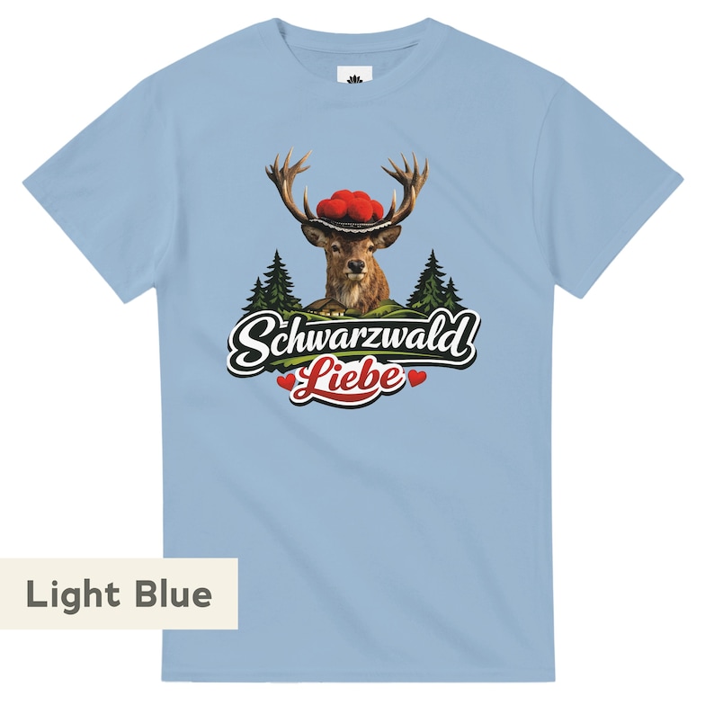 May include: A light blue short-sleeve t-shirt featuring a graphic of a deer wearing a traditional hat with red pom-poms, set against a forest background. Below the deer, the text reads Schwarzwald Liebe with two small red hearts.