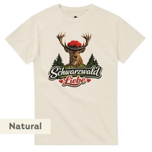 May include: A natural-colored t-shirt featuring a graphic of a deer wearing a hat with red pom-poms, set against a backdrop of pine trees and a small house. Below the deer, the text reads Schwarzwald Liebe with two small red hearts.