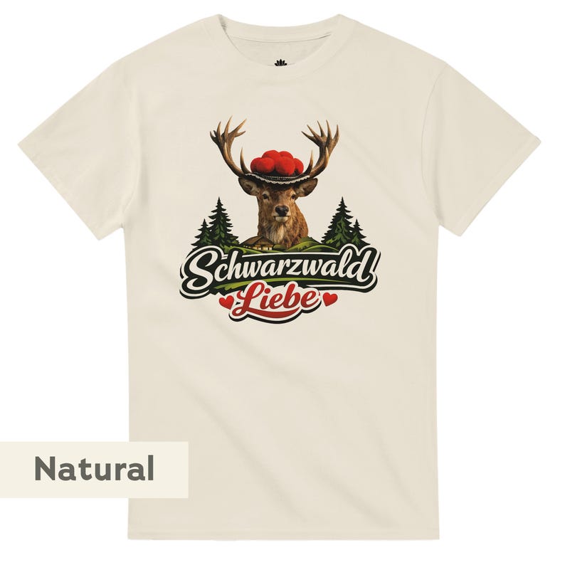 May include: A natural-colored t-shirt featuring a graphic of a deer wearing a hat with red pom-poms, set against a backdrop of pine trees and a small house. Below the deer, the text reads Schwarzwald Liebe with two small red hearts.
