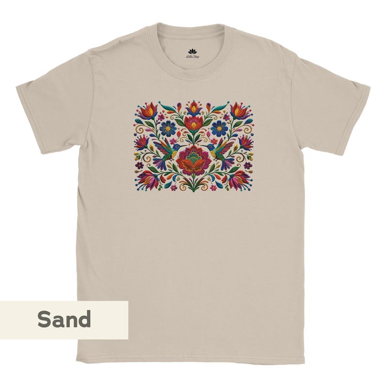 May include: A sand-colored short-sleeve t-shirt featuring a central graphic print of two hummingbirds surrounded by symmetrical, colorful floral patterns in shades of red, blue, green, and yellow. The shirt has a crew neckline and a small logo.