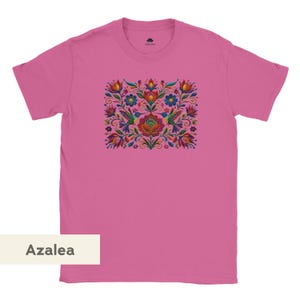 May include: A short-sleeved azalea pink t-shirt featuring a central rectangular graphic print. The design displays a symmetrical arrangement of colorful flowers, swirling foliage, and two hummingbirds in shades of red, blue, and green.