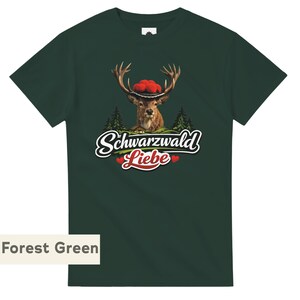 May include: A forest green short-sleeve t-shirt featuring a graphic of a deer wearing a traditional red pom-pom hat. Below the deer are green pine trees and the white text "Schwarzwald" above the red text "Liebe" with two small red hearts.
