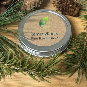 Puede incluir: Un bote plateado de RemedyRoots Pine Resin Salve con una etiqueta marrón y un logotipo de hoja verde. El bote está rodeado de agujas de pino verdes y piñas marrones, sobre una superficie de madera.