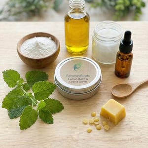 Puede incluir: Una colección de productos naturales para el cuidado de la piel. La imagen muestra un cuenco de madera con polvo blanco, un tarro de cristal, un frasco cuentagotas, una cuchara de madera, una lata de Lemon Balm & Lysine Salve, un cubo de cera amarilla y hojas verdes frescas.