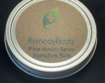 Puede incluir: Una lata redonda plateada de RemedyRoots Pine Resin Salve para pieles sensibles. La etiqueta es marrón con texto verde y blanco y un logotipo de hoja verde. La lata está sobre una superficie negra.