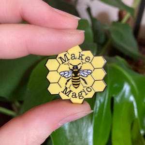 May include: A yellow and black enamel pin in the shape of a honeycomb. The pin features a bee illustration and the words "Make Magic". The pin is held in front of a green leafy background.