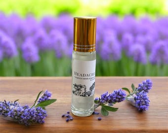 May include: A clear glass roll-on bottle with a gold cap, labelled "Headache." The bottle is on a wooden surface with sprigs of purple lavender. The background features a blurred field of purple flowers.