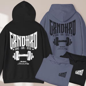 Könnte beinhalten: Schwarze und blaue Kapuzenpullover mit dem Text "GRNDHRD" und einer Hantel-Grafik. Die Kapuzenpullover tragen auch den Text "Grind - Gain - Repeat" und "BUILD NOT BORN". Mehrere gefaltete Kapuzenpullover sind ebenfalls zu sehen.
