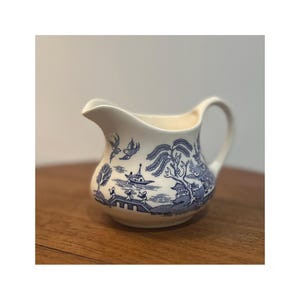 Puede incluir: Cremera de cerámica color crema con un patrón azul de Willow. La cremera presenta un pico, un asa y un diseño de paisaje detallado con árboles, puentes y un barco. Una pieza clásica para servir crema o leche.