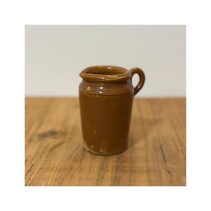 May include: A small, brown ceramic jug with a handle and a spout. The jug has a slightly rough texture and a glossy finish. It sits on a wooden surface.