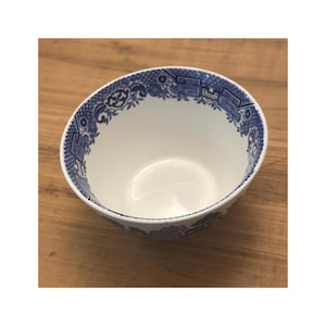 Puede incluir: Un cuenco de cerámica blanca con un patrón floral y arquitectónico azul alrededor del borde. El cuenco está vacío y descansa sobre una superficie de madera.