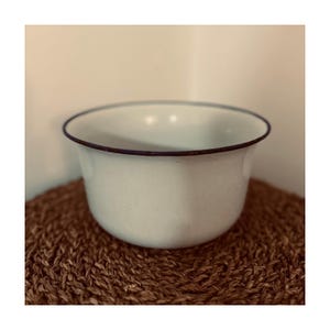 May include: A white enamel bowl with a dark blue rim sits on a woven brown mat. The bowl has a rounded shape and a smooth surface. The rim is a thin, dark blue line that contrasts with the white of the bowl.