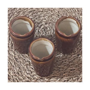 May include: Three small, brown ceramic pots with a textured exterior and a lighter brown rim. The pots are cylindrical with a wide opening and sit on a woven, tan surface. The pots are likely for small plants or storage.